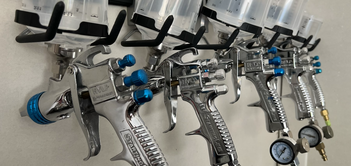 Photo of five Chrome and Blue DeVilbiss Starting Line Paint Guns with 3M PPS 2.0 Disposable Cups on Hooks Hanging on Wall in a Row